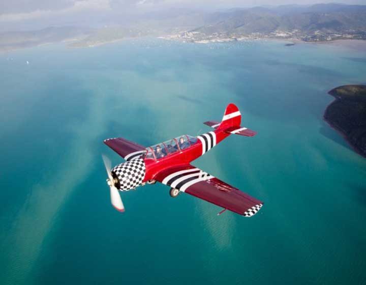 Red and white aerobatic plane with a chequered nose flying over Pioneer Bay with Airlie Beach and the hills behind