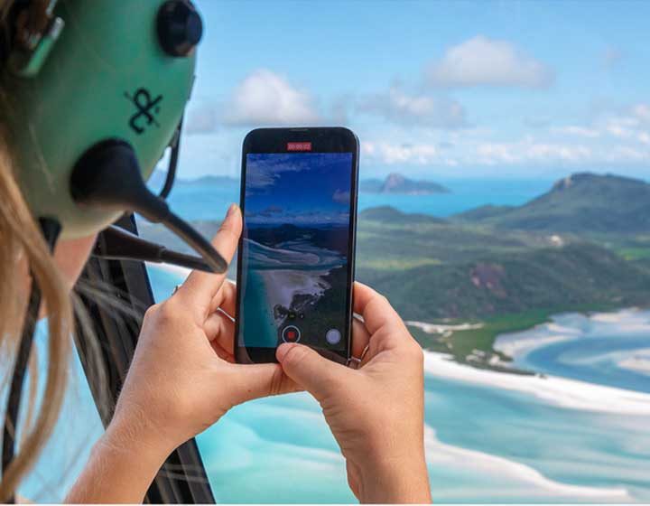 Passenger in a headset filming Hill Inlet and the swirling white sand of Whitehaven Beach on a phone from a helicopter