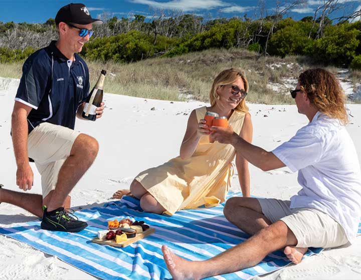 Helicopter pilot pouring bubbles for two guests on a picnic rug on Whitehaven Beach, with a cheese platter on the sand