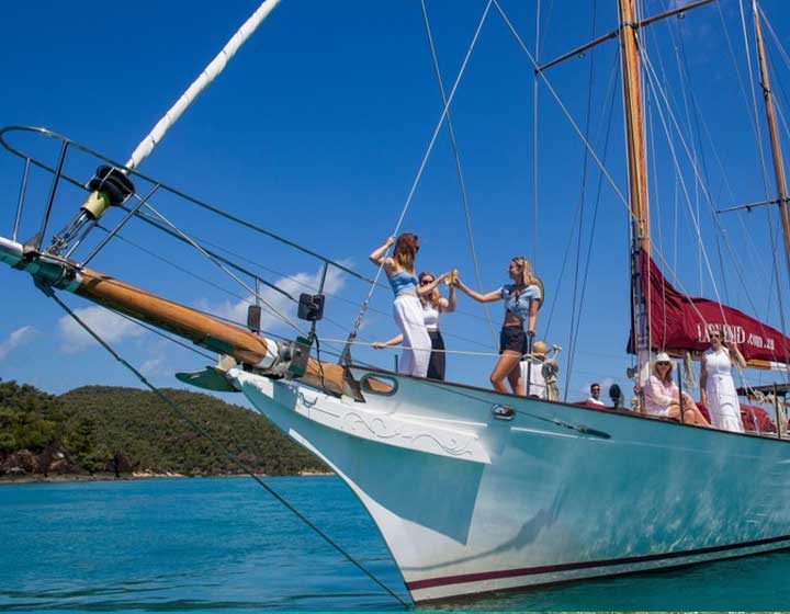 Guests toasting on the bow of Lady Enid, a classic timber sailing yacht run by Iconic Whitsunday Adventures, anchored off a Whitsunday island