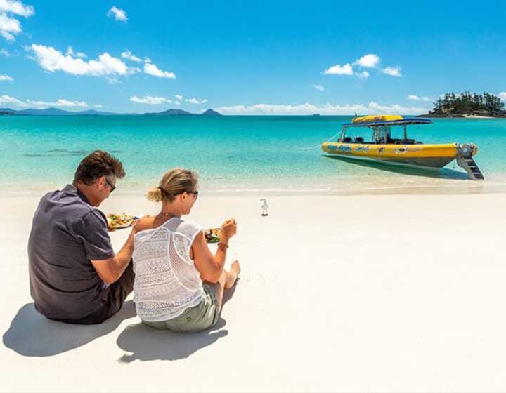 Couple sitting on the white silica sand of Whitehaven Beach with a picnic lunch, an Ocean Rafting boat at the water's edge behind them