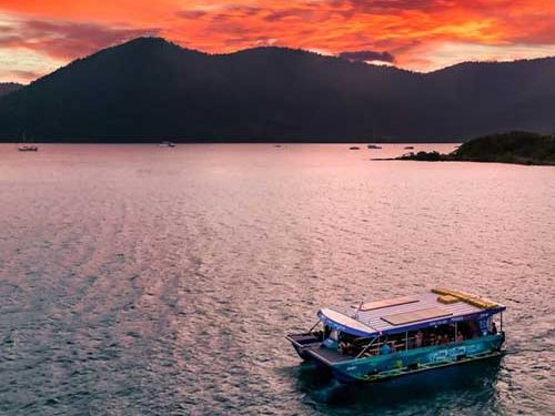 Pioneer Adventures glass bottom boat on calm water near Shute Harbour under a red sunset sky, with the island hills behind