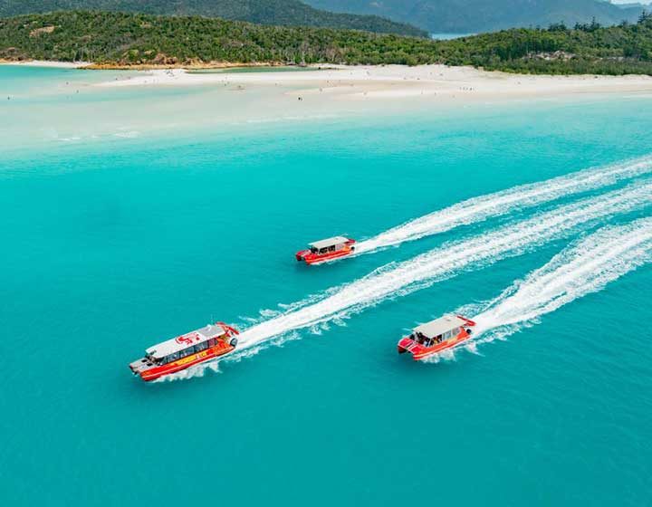 Three red Red Cat Adventures boats speeding across the turquoise water off Hill Inlet, with the white silica sand of Whitsunday Island behind
