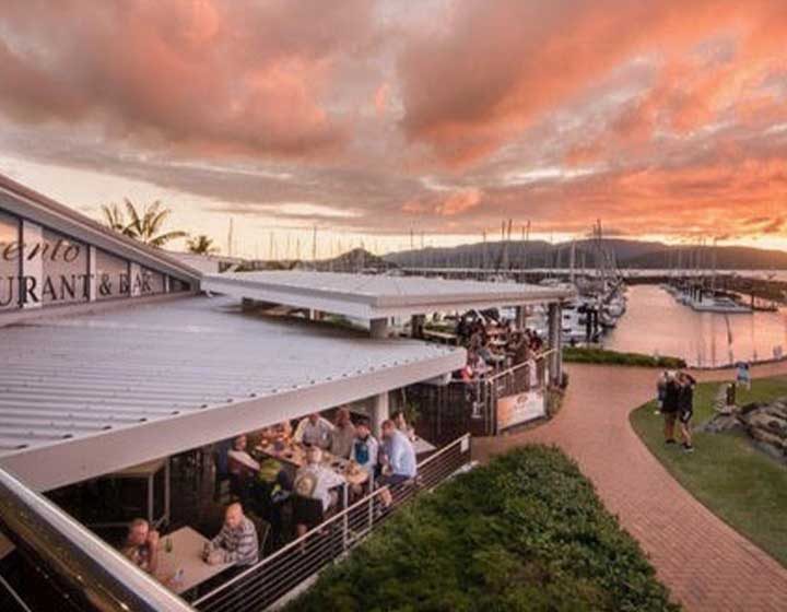 Diners on the deck of Sorrento's Restaurant and Bar at Coral Sea Marina, Airlie Beach, under a pink sunset sky over the moored yachts