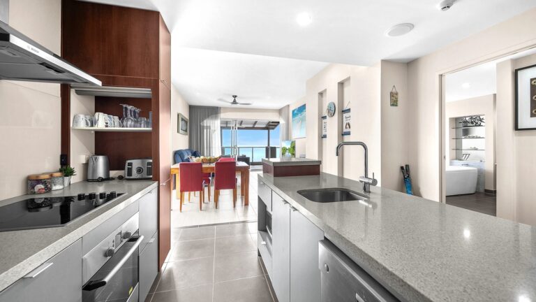 Galley kitchen with Bosch rangehood and oven, stone benches and island sink looking through the dining area to the balcony and Coral Sea, 1 bedroom apartment at Whitsunday Reflections, Airlie Beach