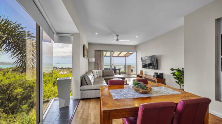 Timber dining table and leather lounge with floor-to-ceiling windows over the hillside and Coral Sea, 1 bedroom apartment at Whitsunday Reflections, Airlie Beach