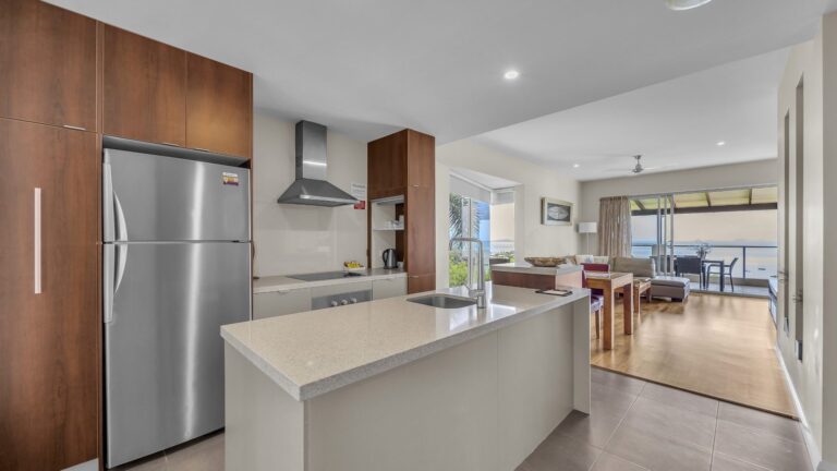 Kitchen with full-size stainless fridge, Bosch rangehood and stone island bench looking through the lounge to the balcony and Coral Sea, 1 bedroom apartment at Whitsunday Reflections, Airlie Beach