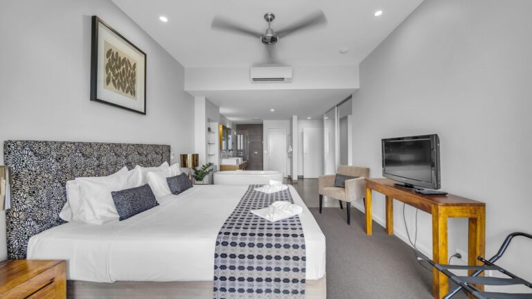 King bed with patterned headboard, TV and armchair looking through to the ensuite with freestanding bath and walk-in shower, 1 bedroom Premium Ocean View apartment at Whitsunday Reflections, Airlie Beach