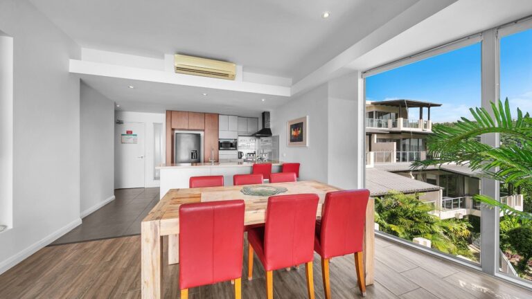 Timber dining table for six beside floor-to-ceiling windows over the hillside gardens, with the kitchen behind, 2 bedroom apartment at Whitsunday Reflections, Airlie Beach