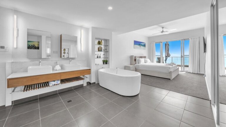 Open-plan main ensuite with freestanding bath and twin basins beside a king bed and sliding doors to a balcony over the Coral Sea, 2 bedroom apartment at Whitsunday Reflections, Airlie Beach