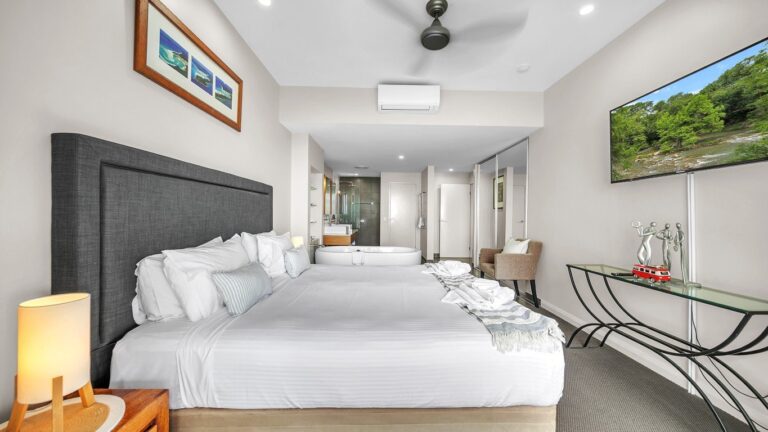 King bed with wall-mounted TV and air conditioning, looking through to the freestanding bath and ensuite, 2 bedroom apartment at Whitsunday Reflections, Airlie Beach