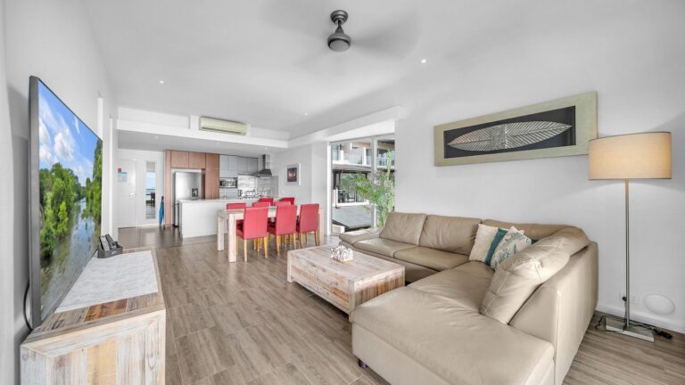 Open-plan lounge with leather corner sofa, large Smart TV and floor lamp, dining and kitchen beyond, 2 bedroom apartment at Whitsunday Reflections, Airlie Beach