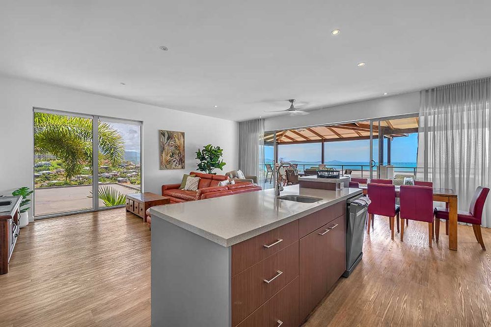 Open-plan kitchen with stone island bench, lounge and dining opening to a covered deck with Coral Sea views, 3 bedroom apartment at Whitsunday Reflections, Airlie Beach