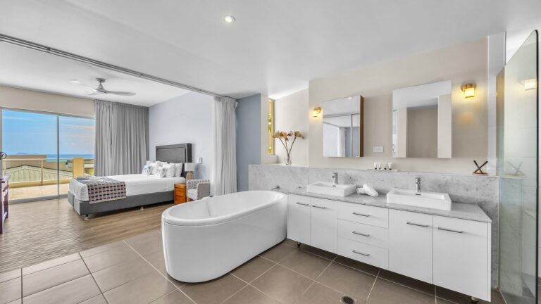 Open-plan main bedroom with freestanding bath and twin basins on a marble vanity beside a king bed and sliding doors to the balcony and Coral Sea, 3 bedroom apartment at Whitsunday Reflections, Airlie Beach