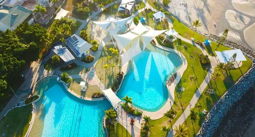 Aerial view of Airlie Beach Lagoon on the foreshore, with shade sails over the pools, lap lanes, palm-lined lawns and the rock wall to the sea