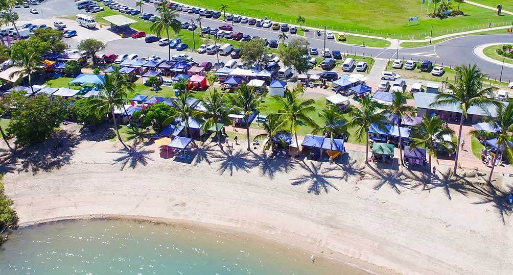 Aerial view of Saturday market stalls under the palms on the Airlie Beach foreshore, beside the beach