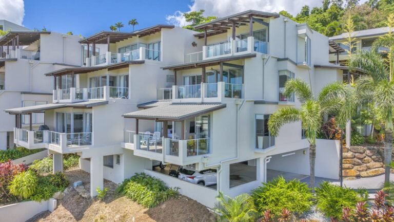 White apartment buildings with glass balconies and timber pergolas above tropical gardens and a rock retaining wall, Whitsunday Reflections, Airlie Beach
