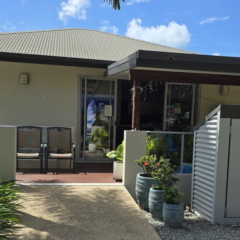 Paved path leading up to the Whitsunday Reflections reception door, with Reception lettering on the wall, a covered deck with chairs and potted plants, Airlie Beach