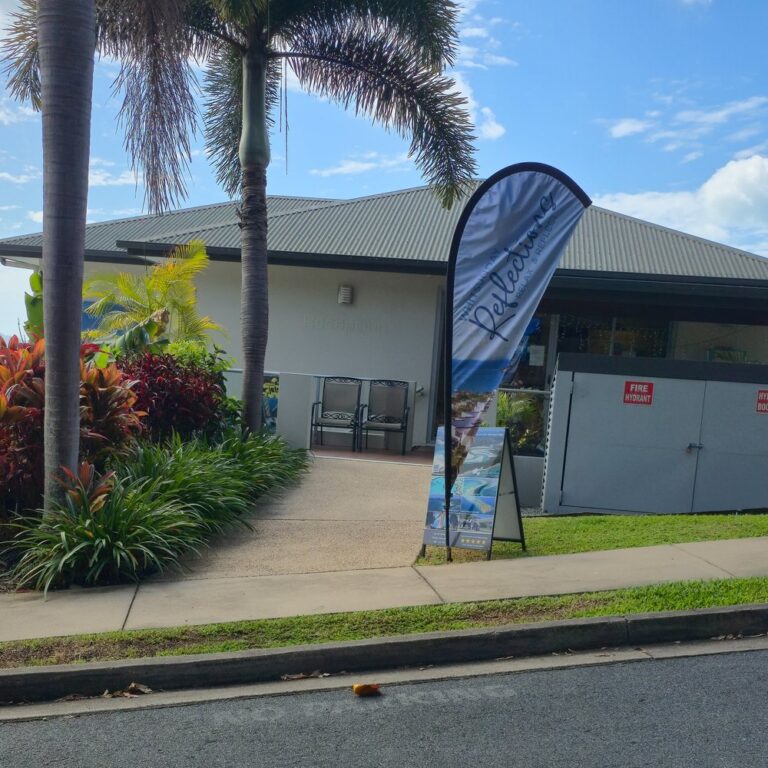 Whitsunday Reflections reception building seen from the street, with palms, tropical gardens and a Whitsunday Reflections flag at the start of the path, Airlie Beach