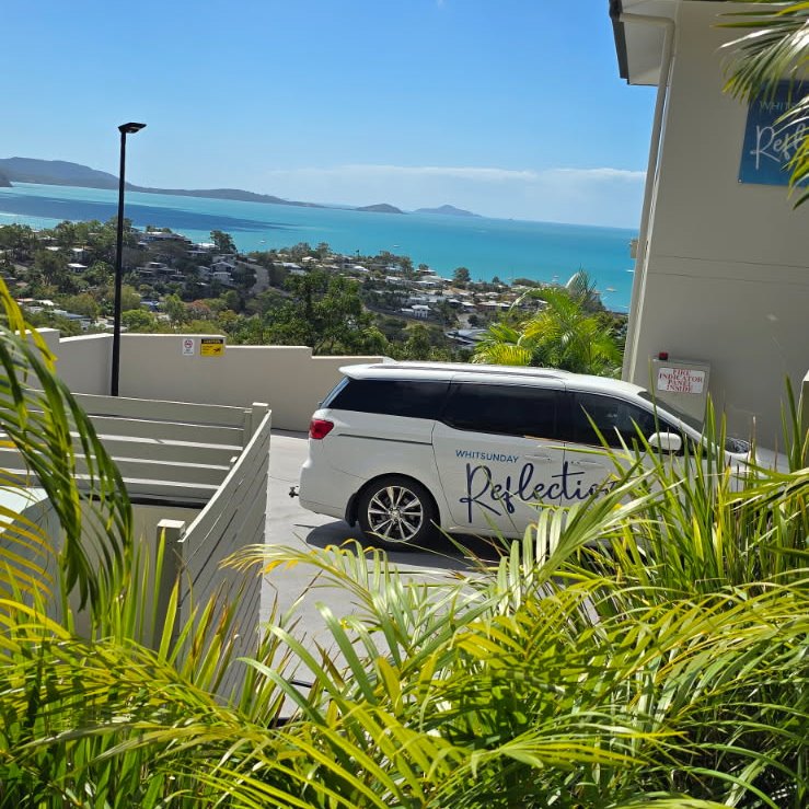 Car park beside reception with the Whitsunday Reflections van and a view over Airlie Beach to the Coral Sea and Whitsunday Islands, framed by palms
