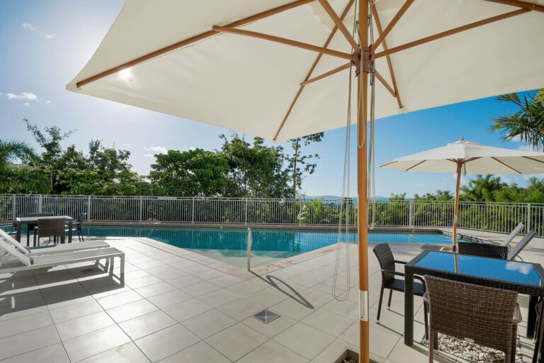 Shaded pool deck with market umbrellas, wicker dining settings and sun lounges beside the lap pool, with a glimpse of the Coral Sea through the trees, Whitsunday Reflections, Airlie Beach