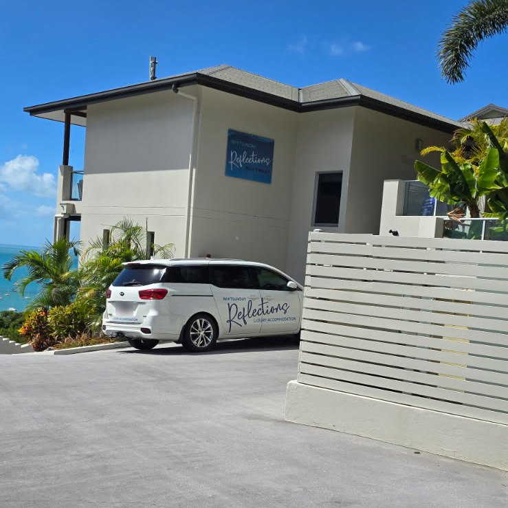 Whitsunday Reflections reception building with its blue sign and the white Whitsunday Reflections van parked out front, glimpse of the Coral Sea at left, Airlie Beach