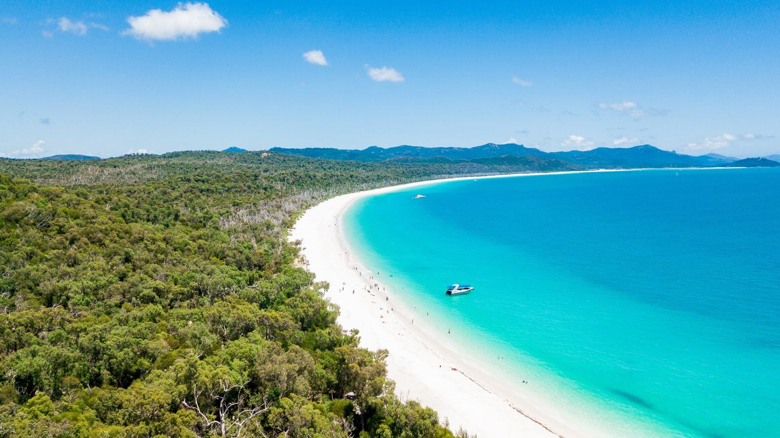 Aerial view of Whitehaven Beach curving along Whitsunday Island, with a boat anchored off the white silica sand and swimmers in the shallows