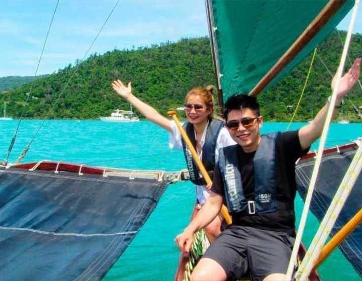 Couple in life jackets waving from a sailing outrigger canoe on turquoise water off Airlie Beach