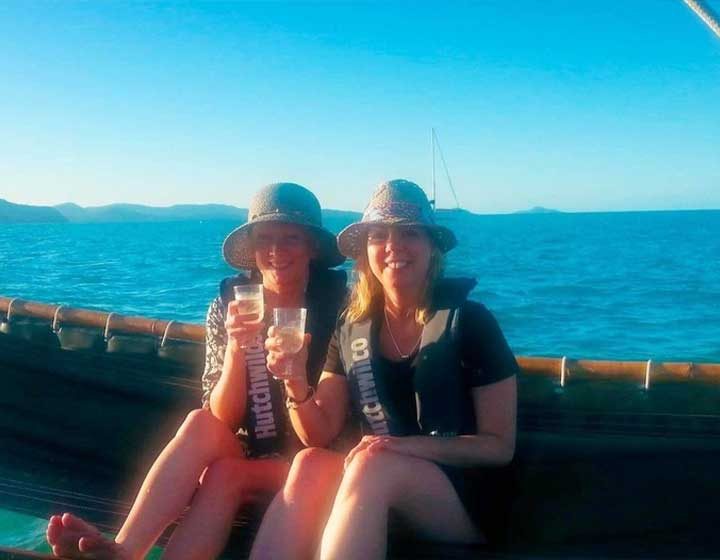 Two women in hats raising glasses of bubbles on a sailing outrigger canoe off Airlie Beach, with the islands behind
