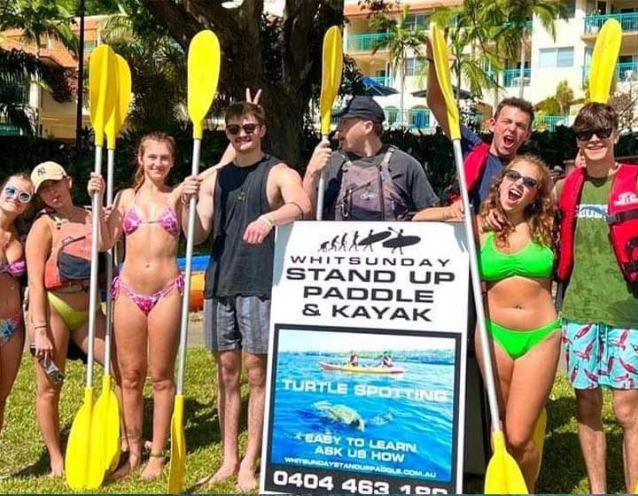 Group of young paddlers with yellow kayak paddles around the Whitsunday Stand Up Paddle and Kayak sign on the Airlie Beach foreshore