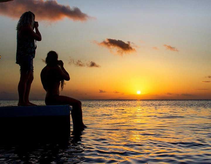 Two guests silhouetted on the stern of a sailing catamaran as the sun sets over the Whitsundays